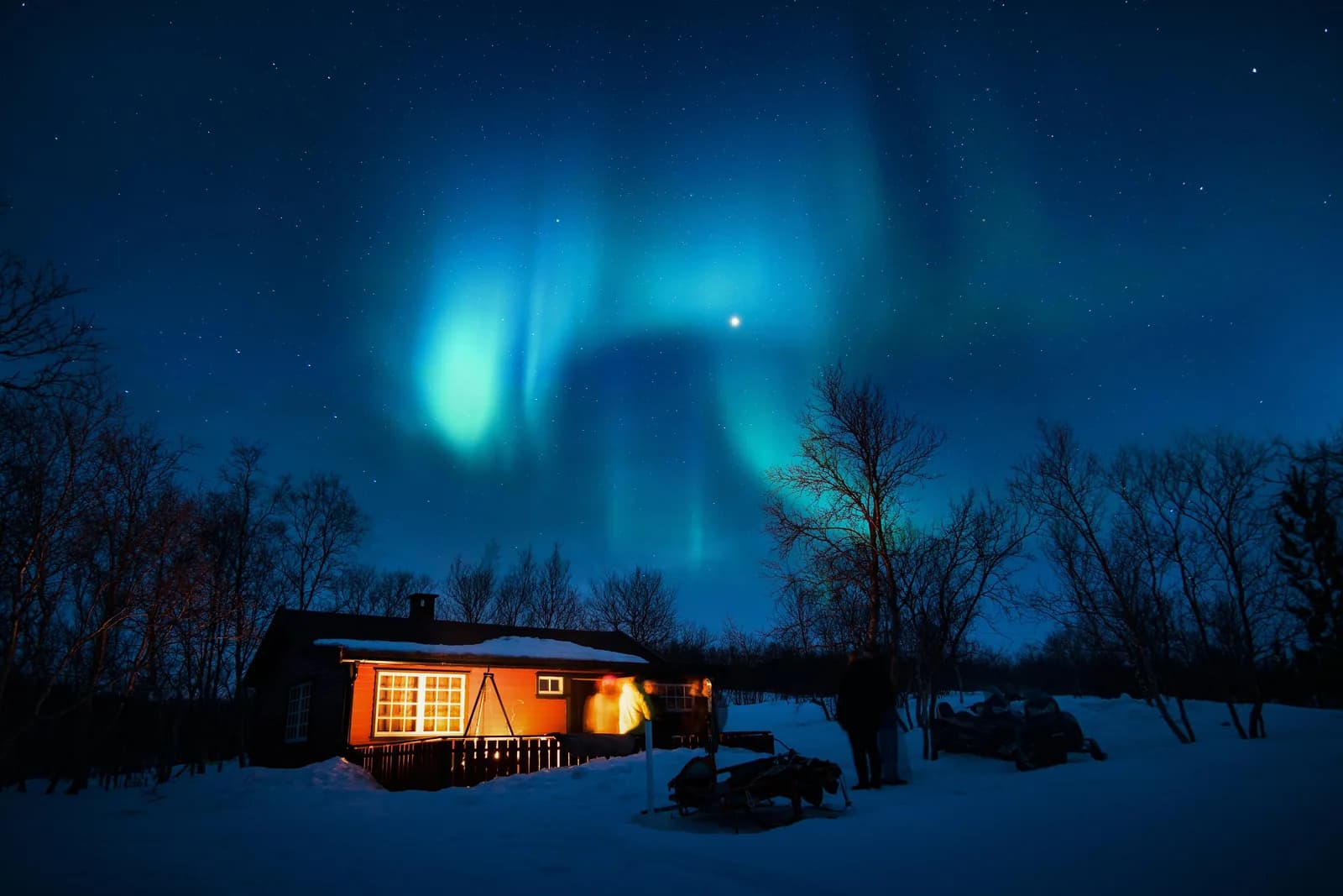Aurore boréale verte et turquoise au-dessus d’un chalet éclairé dans la neige, Finnmark