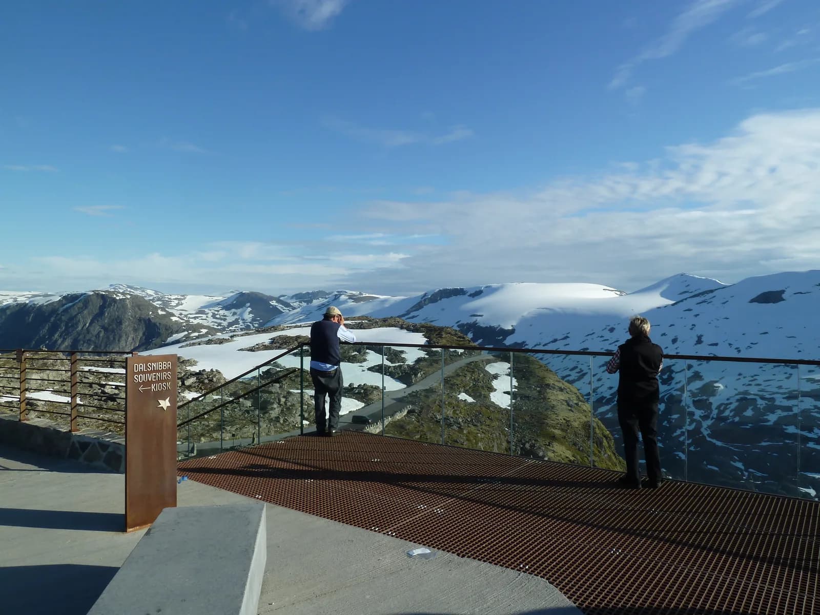 Plateforme panoramique à garde-corps de verre du Geiranger Skywalk au sommet de Dalsnibba, avec des visiteurs face aux montagnes enneigées